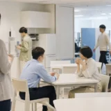職場の休憩スペースで談笑の輪から少し離れて立つ後ろ姿の人物