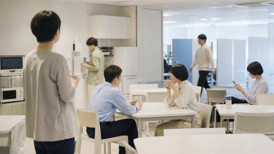 職場の休憩スペースで談笑の輪から少し離れて立つ後ろ姿の人物